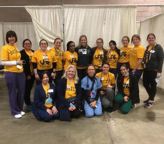 A group of standing and kneeling nurse practitioner alumni, faculty and students wearing gold Drexel t-shirts and scrubs bottoms.